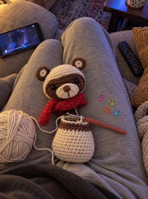 Gehäkelter Bär mit rotem Schal in Arbeit, mit Garn und Häkelnadel auf dem Sofa